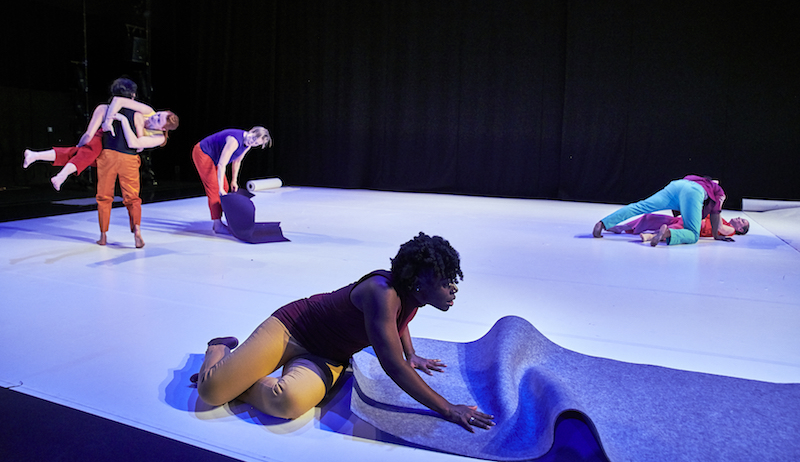 A woman sits in the foreground and rolls out a purple mat. Couples in the background pick one another up or lean over one another. One woman rolls up a mat.